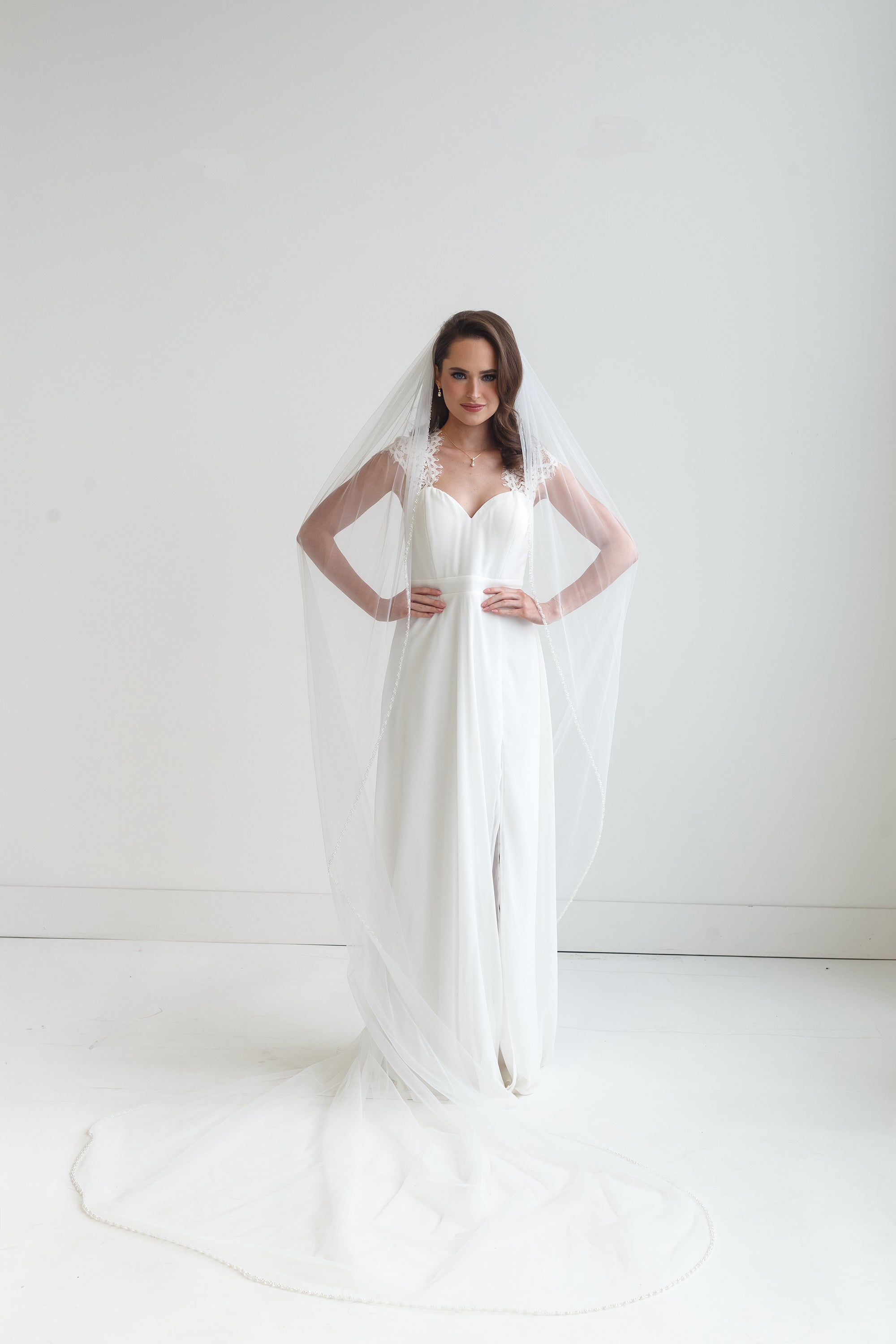 A woman in a white wedding dress stands with her hands on her hips, wearing a long white veil that extends behind her.