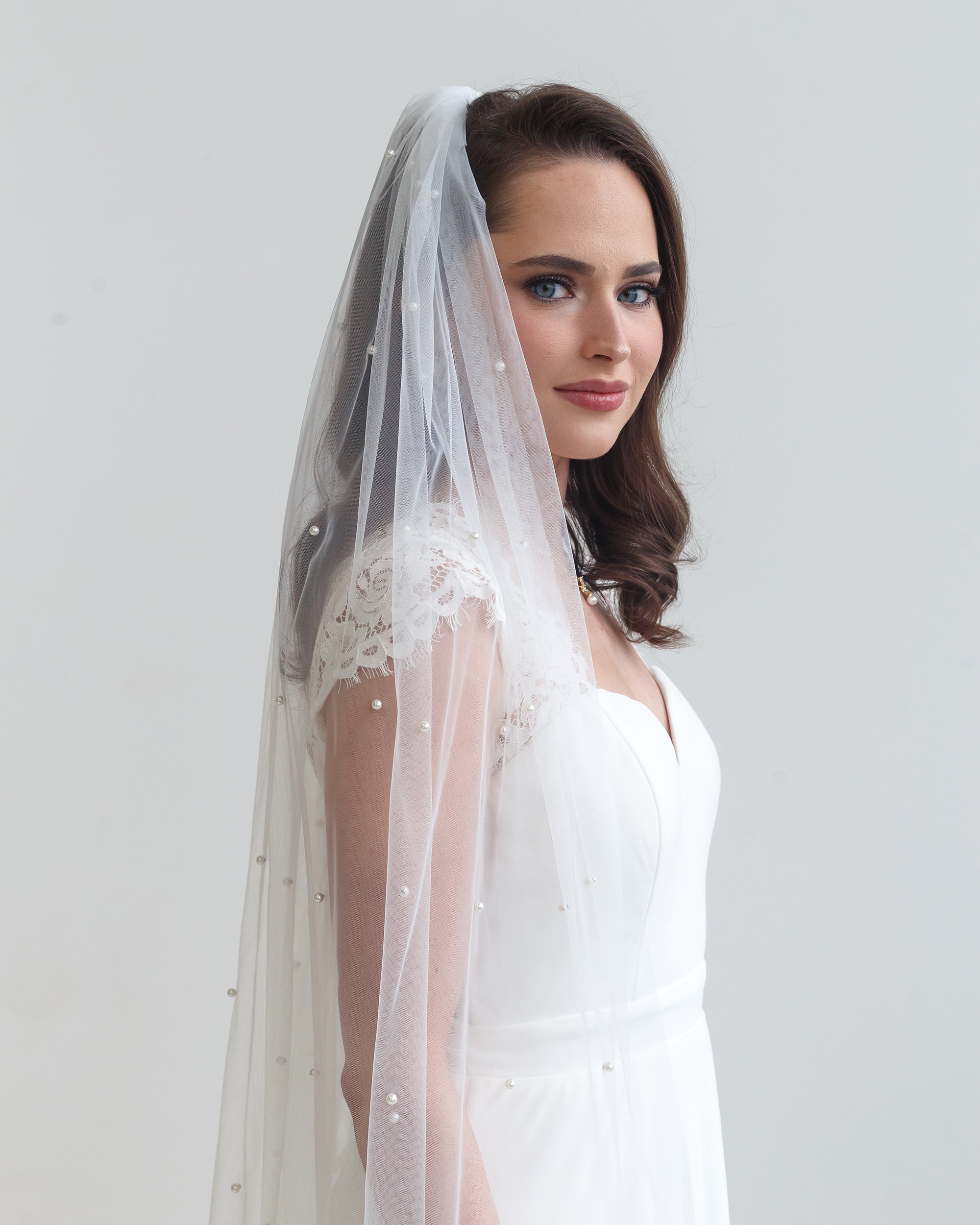 A woman in a white wedding dress with a long veil stands against a plain white background, gazing off to the side.