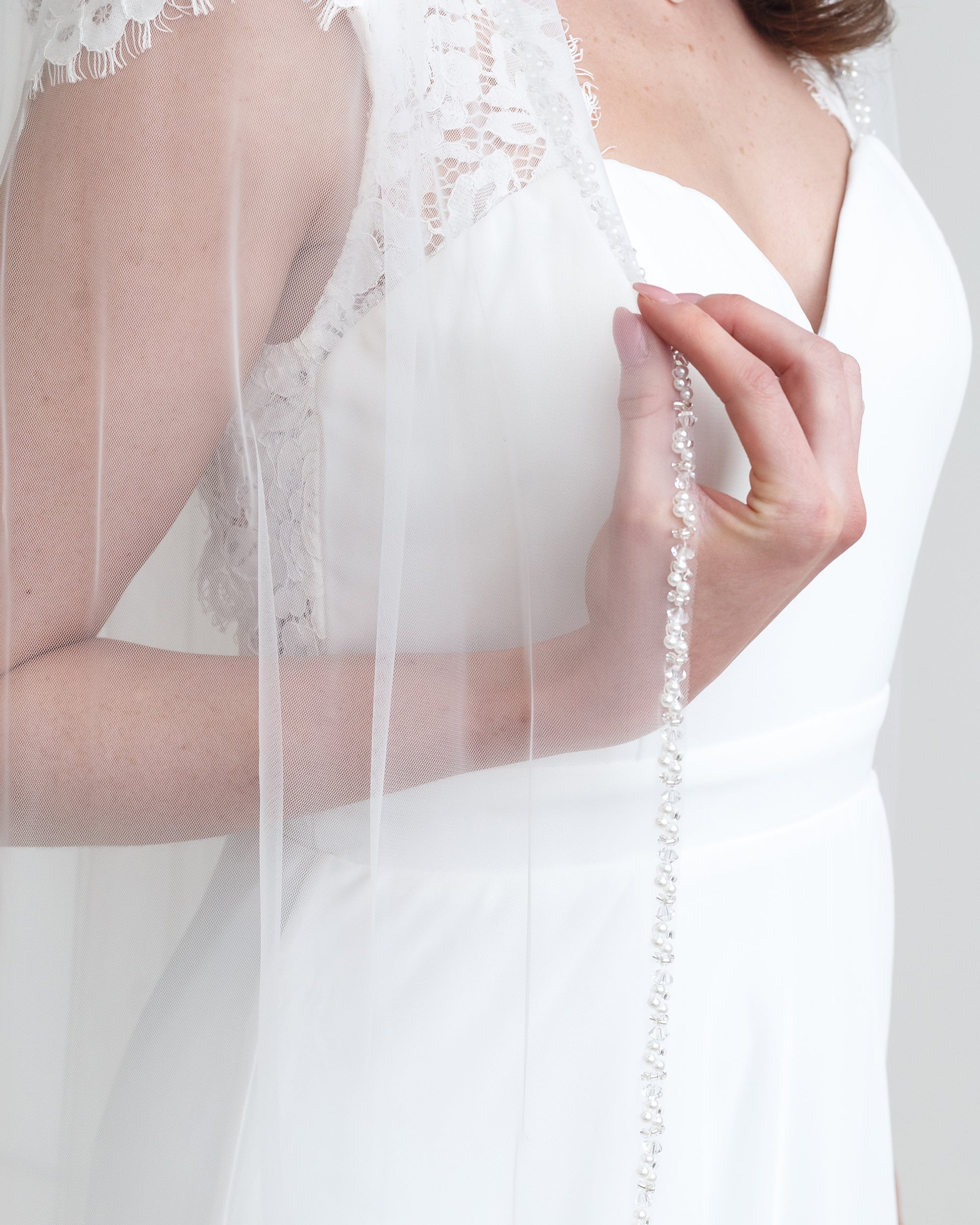 A close-up of a person wearing a white wedding dress with a veil, holding the veil with their hand.