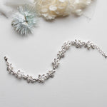A delicate silver bracelet adorned with white pearls and silver leaves rests on a white surface, accompanied by a small white flower and a piece of white fabric.