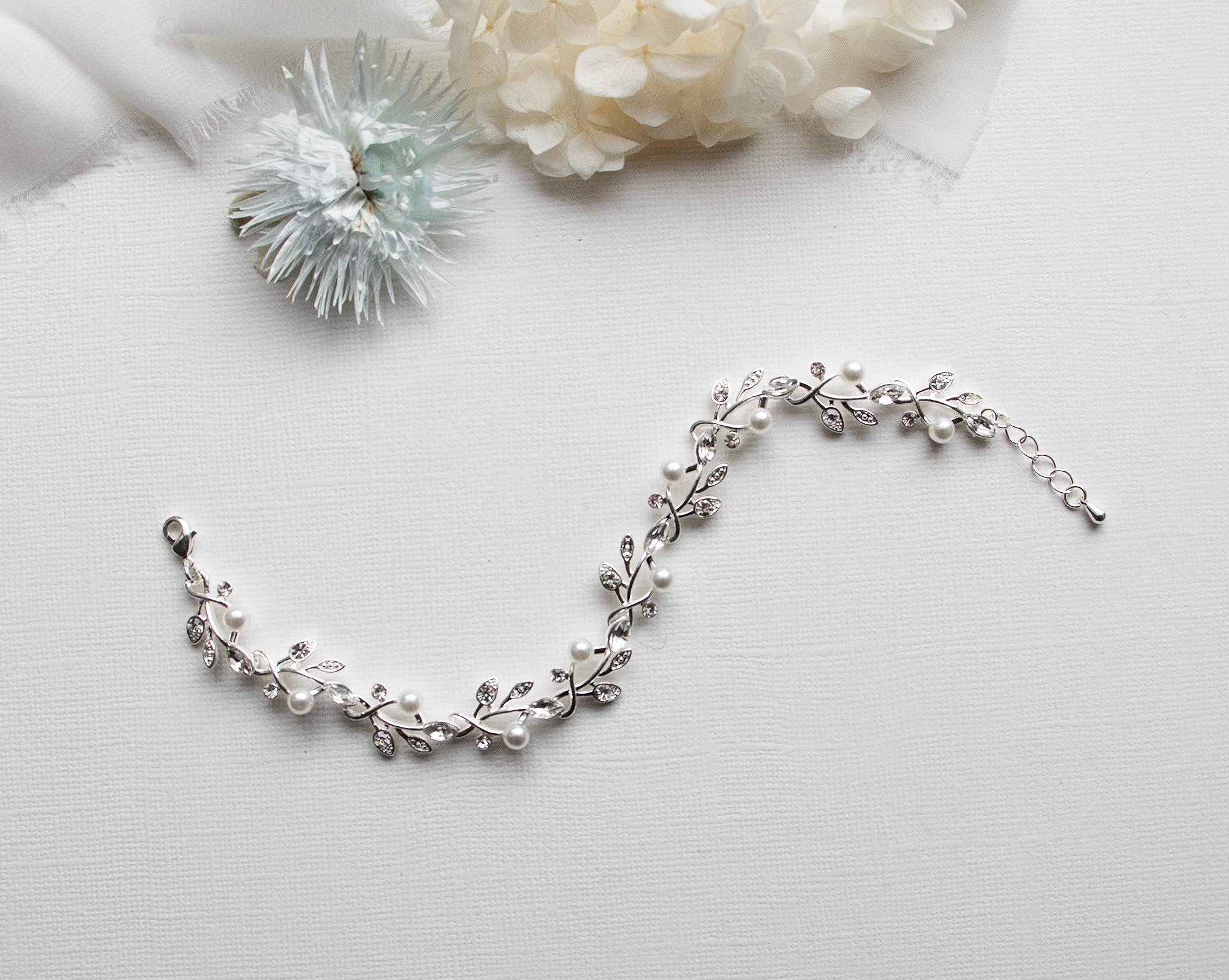 A delicate silver bracelet adorned with white pearls and silver leaves rests on a white surface, accompanied by a small white flower and a piece of white fabric.