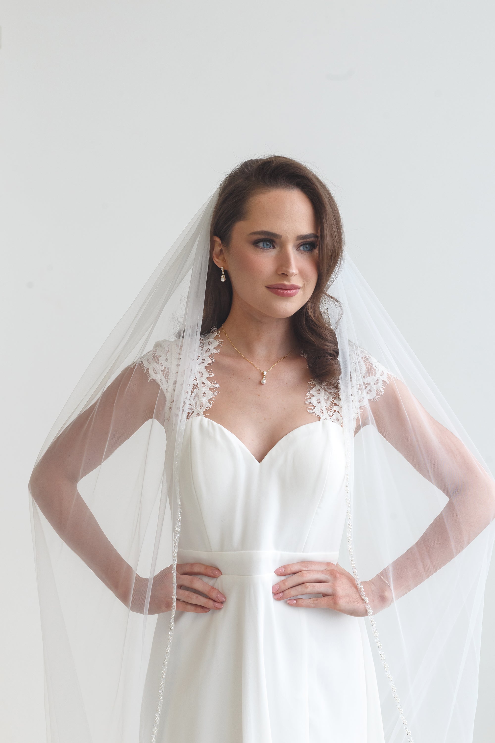A woman in a white wedding dress with a veil stands confidently with her hands on her hips, gazing directly at the camera.