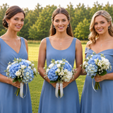 Three women in blue dresses holding bouquets of white and blue flowers stand together in a grassy field.