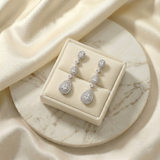 A pair of elegant diamond earrings is displayed in a white jewelry box on a marble surface.
