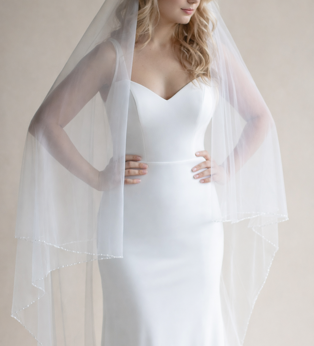 A woman in a white wedding dress with a long veil stands against a beige background, her hands resting on her hips.