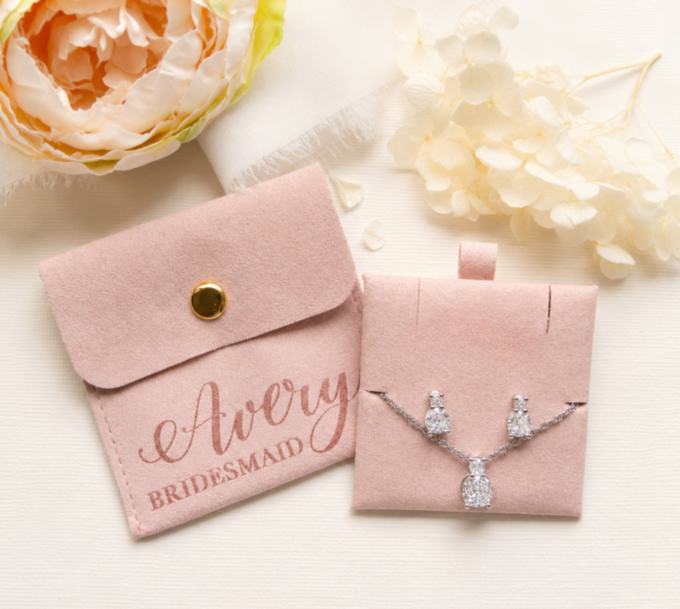 A pink bridesmaid jewelry set, including a necklace and earrings, is displayed alongside a pink satin pouch with the name "Avery" printed on it.