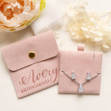 A pink bridesmaid jewelry set, including a necklace and earrings, is displayed alongside a pink satin pouch with the name "Avery" printed on it.