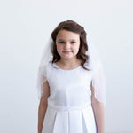 A young girl in a white dress and veil stands against a white background, smiling at the camera.