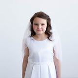 A young girl in a white dress and veil stands against a white background, smiling at the camera.