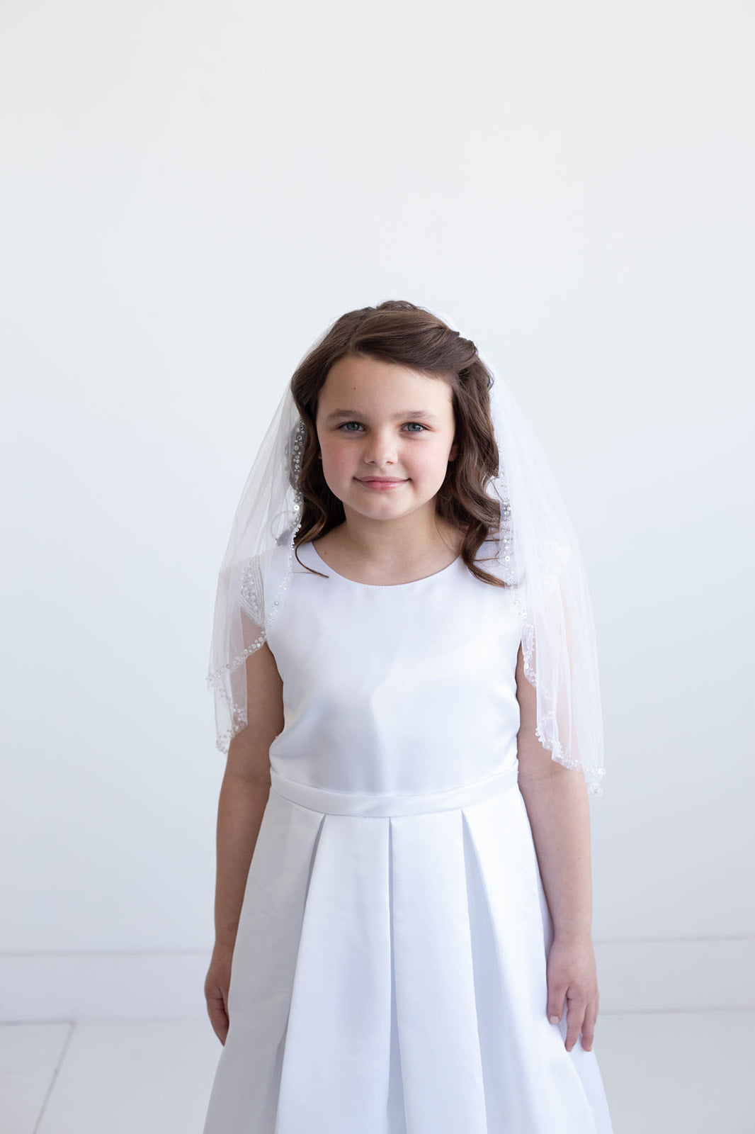 A young girl in a white dress and veil stands against a white background, smiling at the camera.