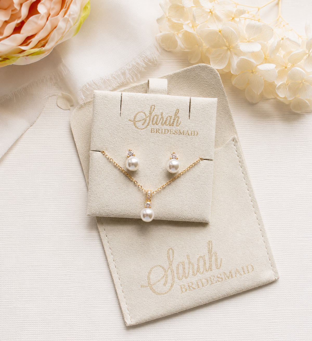 A beige jewelry box with a pearl necklace and earrings, along with a matching beige pouch, sits on a white surface. A pink flower is visible in the background, and the text "Sarah Bridesmaid" is printed on the jewelry box and pouch.