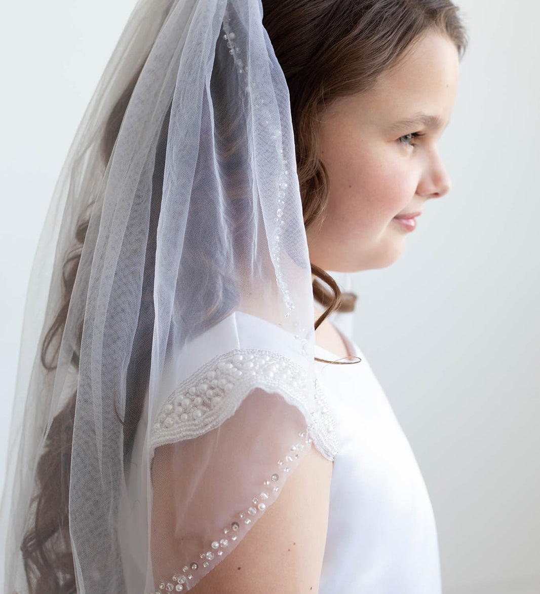 A young girl wearing a white dress with a veil and beaded accents, standing against a plain white background.