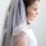 A young girl wearing a white dress with a veil and beaded accents, standing against a plain white background.