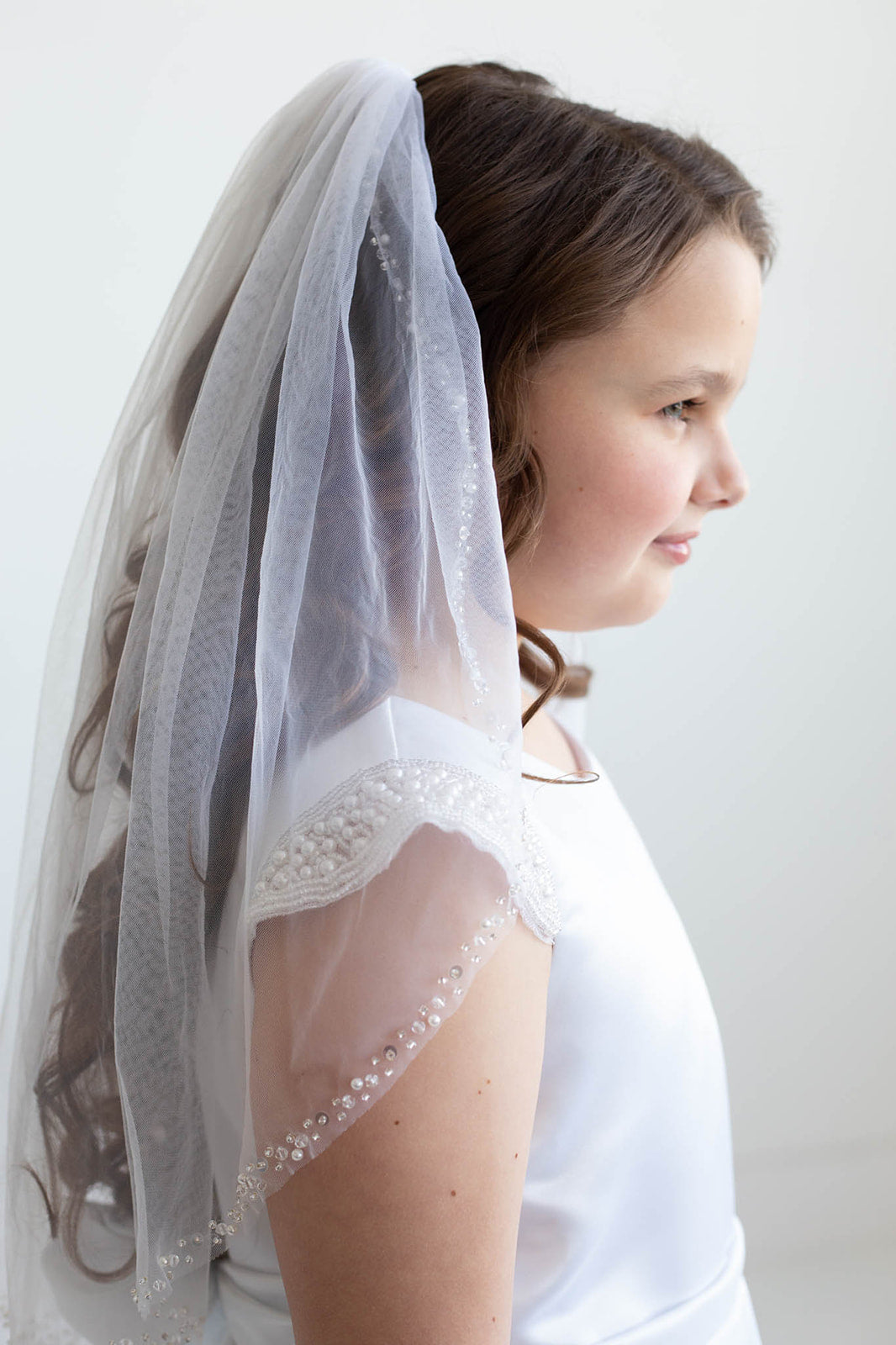 A young girl wearing a white dress with a veil and beaded accents, standing against a plain white background.
