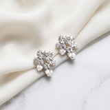 A pair of elegant diamond earrings with large, clear gemstones are displayed on a white fabric surface.