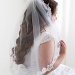 A young girl in a white wedding dress with a veil stands in front of a white wall, her hair styled in curls.