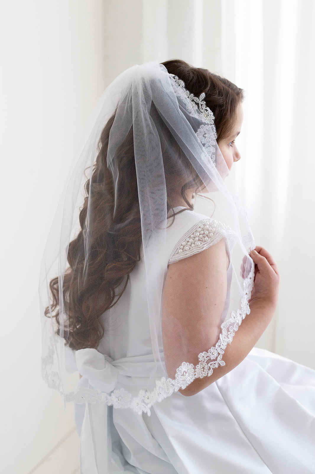 A young girl in a white wedding dress with a veil stands in front of a white wall, her hair styled in curls.