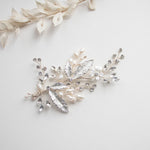 a decorative piece of jewelry with a combination of silver, white, and clear gemstones, including pearls and leaves, arranged on a white background.