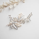 a decorative piece of jewelry with a combination of silver, white, and clear gemstones, including pearls and leaves, arranged on a white background.
