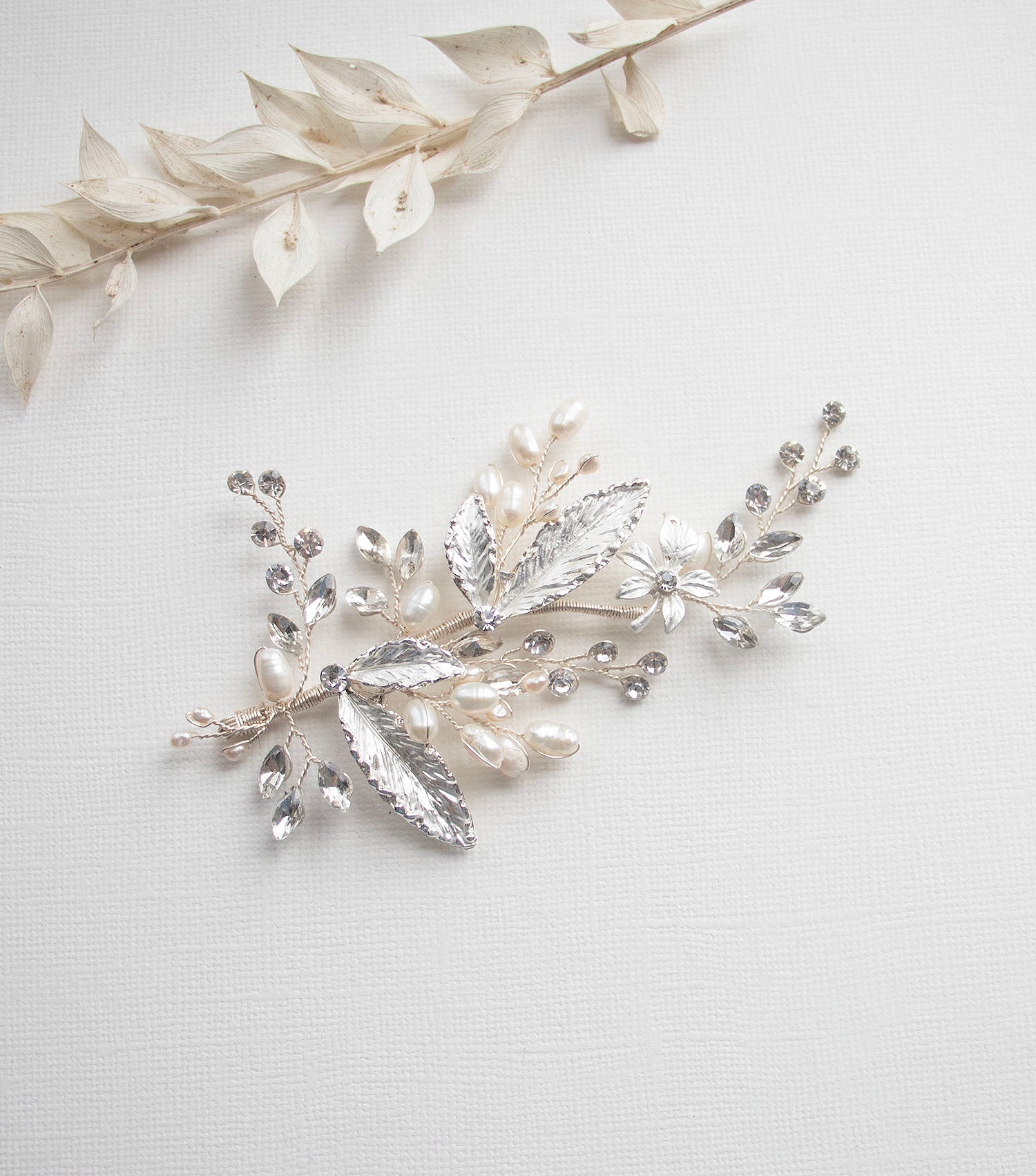 a decorative piece of jewelry with a combination of silver, white, and clear gemstones, including pearls and leaves, arranged on a white background.