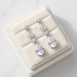 A pair of elegant earrings with large, clear gemstones, displayed in a white jewelry box on a marble surface.