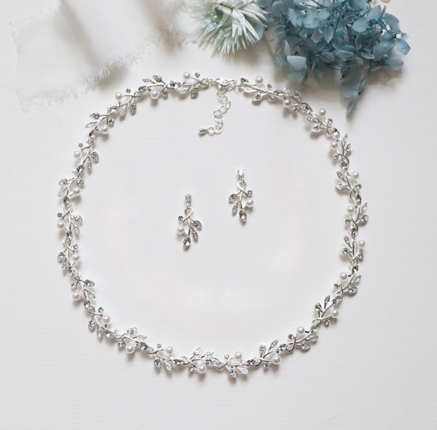 A delicate necklace with a leaf-shaped pendant, earrings, and a bracelet, all adorned with small white pearls and silver leaves, arranged on a white surface.