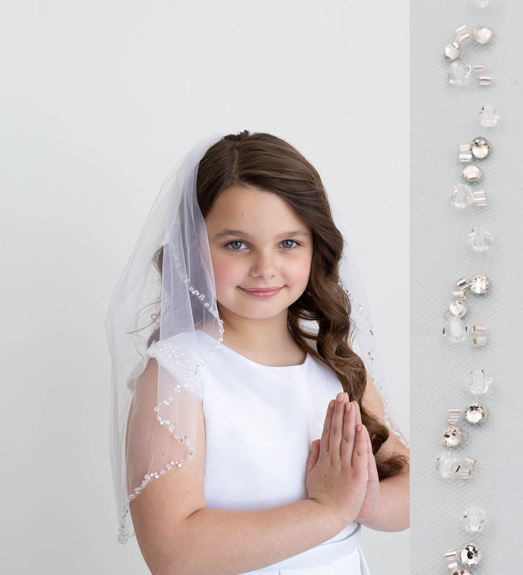 A young girl in a white dress and veil stands against a white background, smiling at the camera.