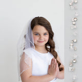 A young girl in a white dress and veil stands against a white background, smiling at the camera.