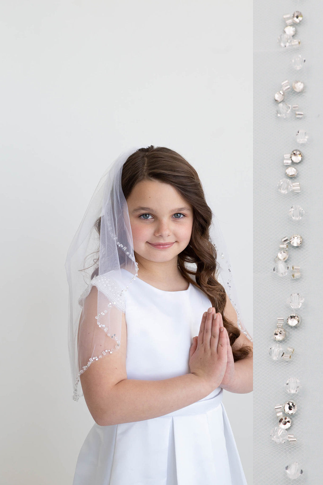 A young girl in a white dress and veil stands against a white background, smiling at the camera.