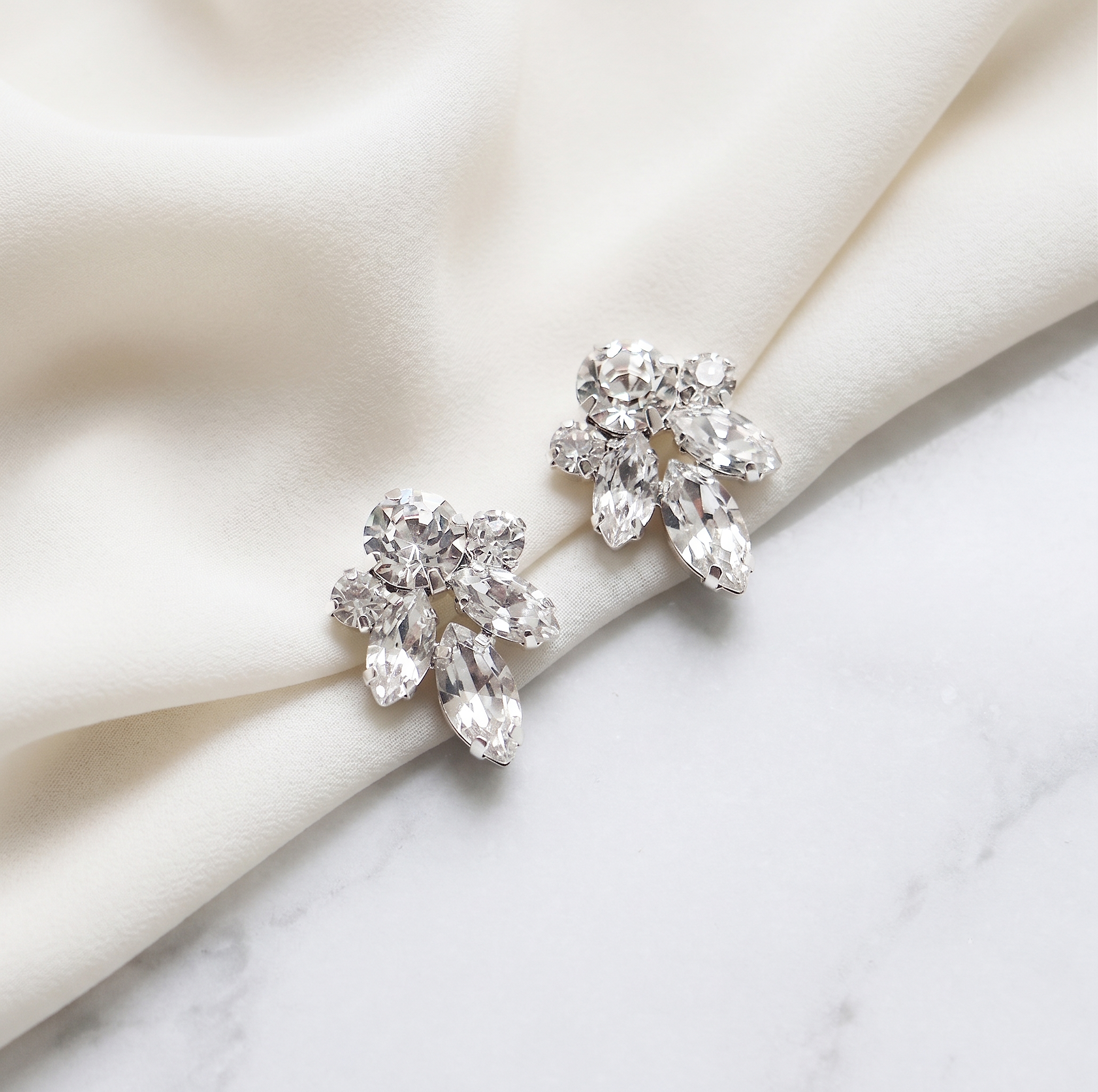 A pair of elegant diamond earrings with large, clear gemstones are displayed on a white fabric surface.