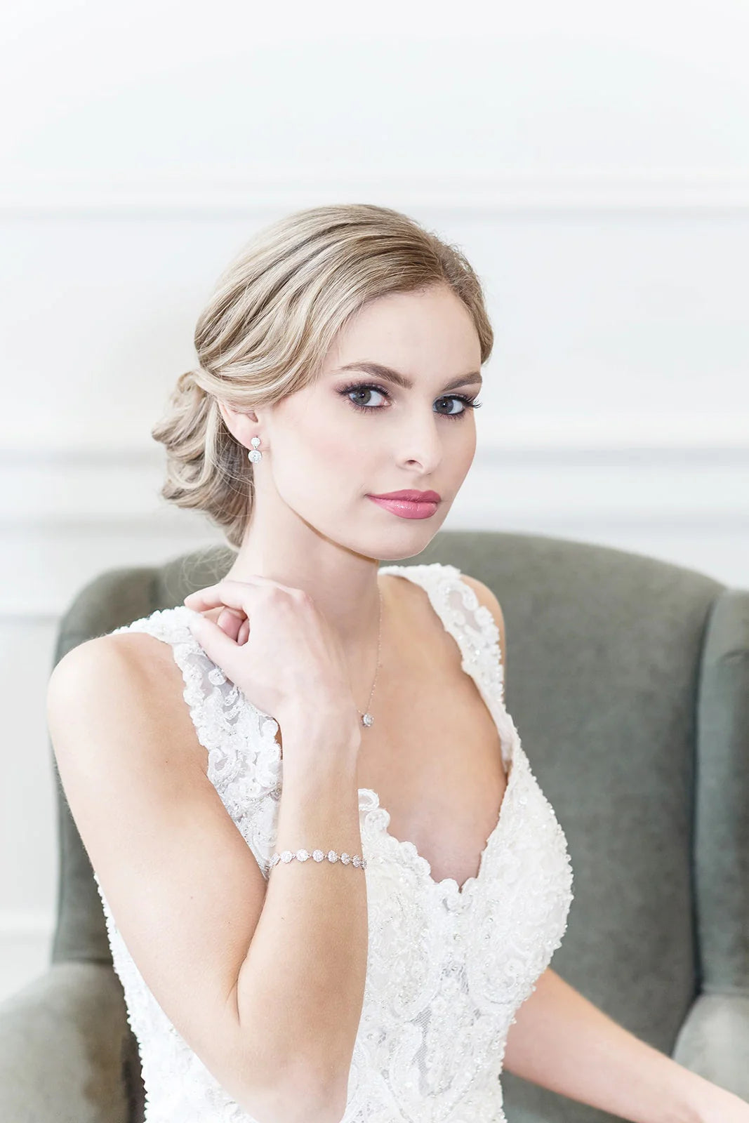 A woman in a white lace wedding dress sits in a chair, her gaze directed off to the side.