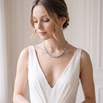 A woman in a white dress with a necklace and earrings stands in front of a white wall, her gaze directed downwards.