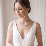A woman in a white dress with a necklace and earrings stands in front of a white wall, her gaze directed downwards.