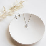 A pair of silver cross earrings and a silver necklace with a cross pendant are displayed on a white plate, accompanied by a delicate white flower.