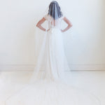 A woman in a white wedding dress stands with her hands on her hips, wearing a long white veil that extends behind her.