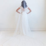 A woman in a white wedding dress stands with her hands on her hips, wearing a long white veil that extends behind her.