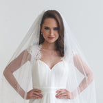 A woman in a white wedding dress with a veil stands confidently with her hands on her hips, gazing directly at the camera.