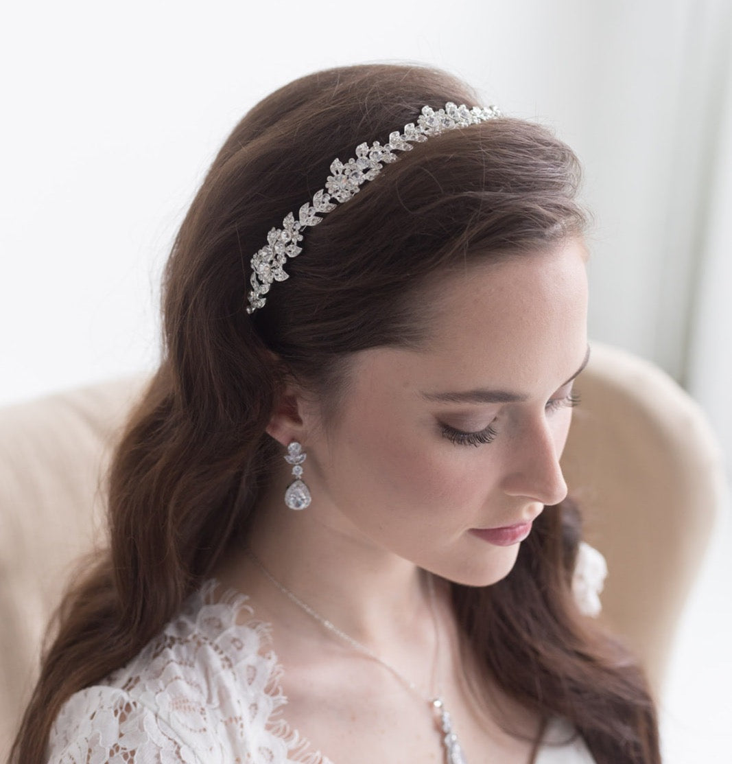 A woman with long brown hair wearing a white lace dress and a decorative headpiece, looking down at her hands.