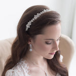 A woman with long brown hair wearing a white lace dress and a decorative headpiece, looking down at her hands.