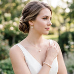A woman in a white dress stands in a garden, wearing pearl earrings and a bracelet.