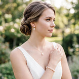 A woman in a white dress stands in a garden, wearing pearl earrings and a bracelet.