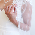 A close-up of a person wearing a white wedding dress with a lace veil, holding the veil with their hand.