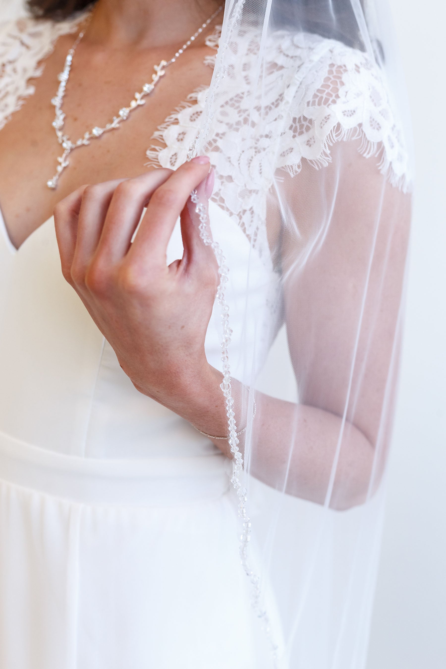 A close-up of a person wearing a white wedding dress with a lace veil, holding the veil with their hand.
