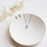 A pair of silver cross earrings and a silver necklace with a cross pendant are displayed on a white plate, accompanied by a delicate white flower.