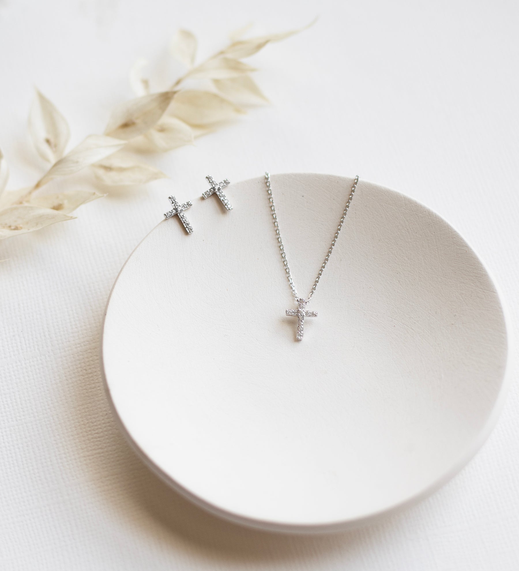 A pair of silver cross earrings and a silver necklace with a cross pendant are displayed on a white plate, accompanied by a delicate white flower.