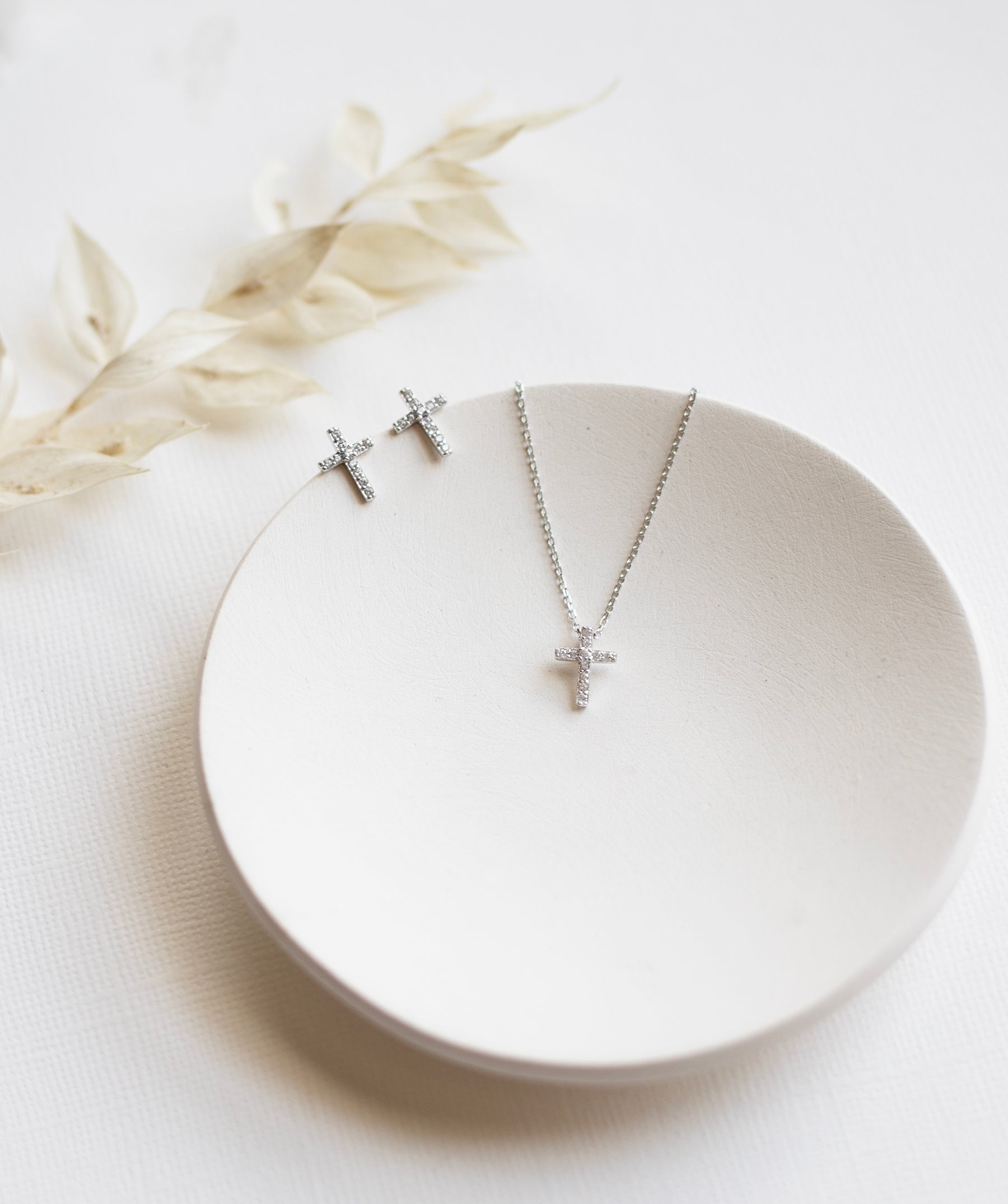 A pair of silver cross earrings and a silver necklace with a cross pendant are displayed on a white plate, accompanied by a delicate white flower.