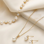 A delicate gold necklace with a row of pearls, a matching gold bracelet, and a pair of pearl earrings are displayed on a white fabric background.