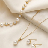 A delicate gold necklace with a row of pearls, a matching gold bracelet, and a pair of pearl earrings are displayed on a white fabric background.