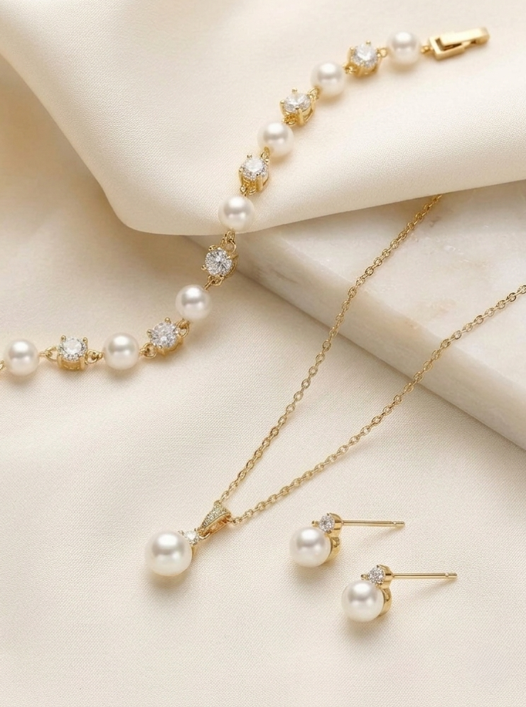 A delicate gold necklace with a row of pearls, a matching gold bracelet, and a pair of pearl earrings are displayed on a white fabric background.