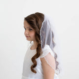 A young girl with long, curly hair wearing a white dress and a veil stands against a plain white background.